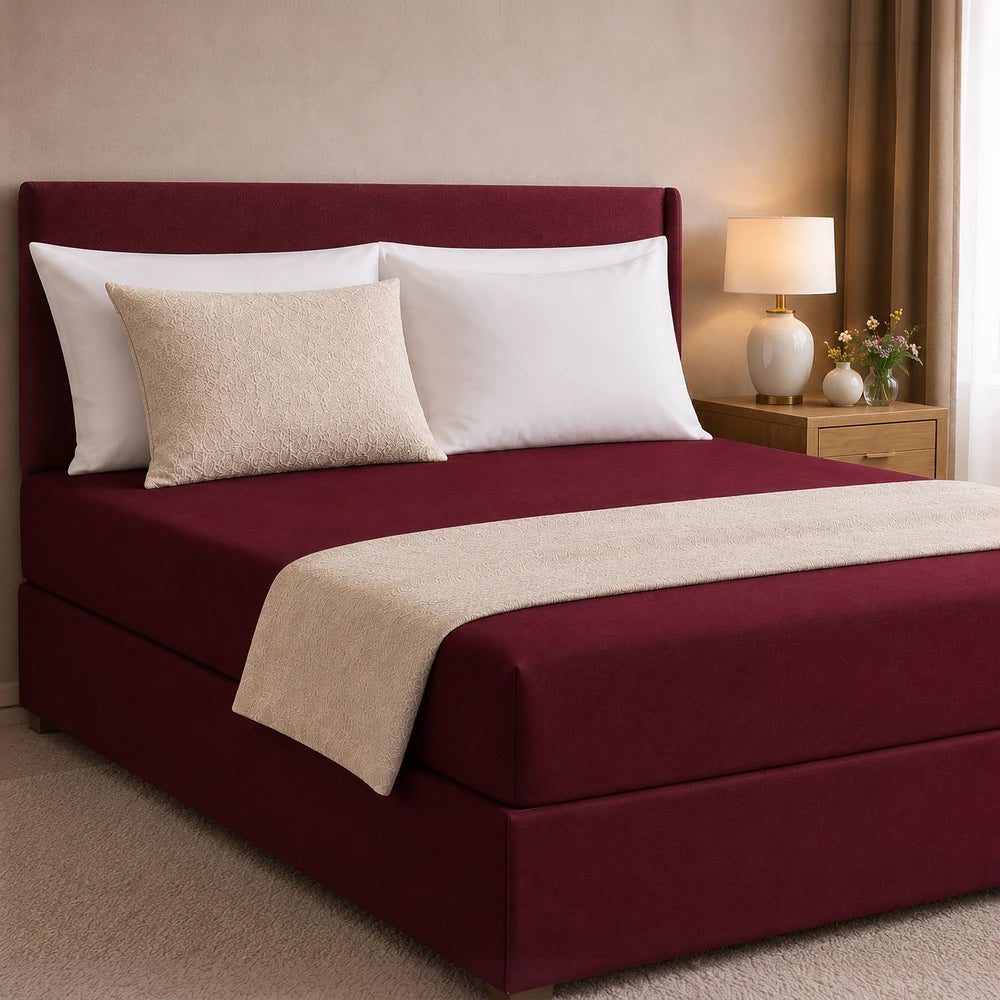 All-in-one fitted sheet and base cover in burgundy with beige quilted accents on a bed with white pillows.