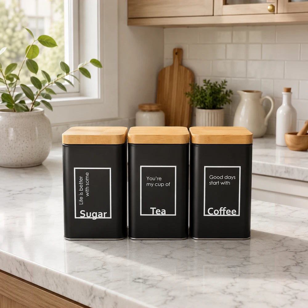 Matte black and bamboo kitchen storage tins trio with labels for Sugar, Tea, and Coffee on a marble countertop.