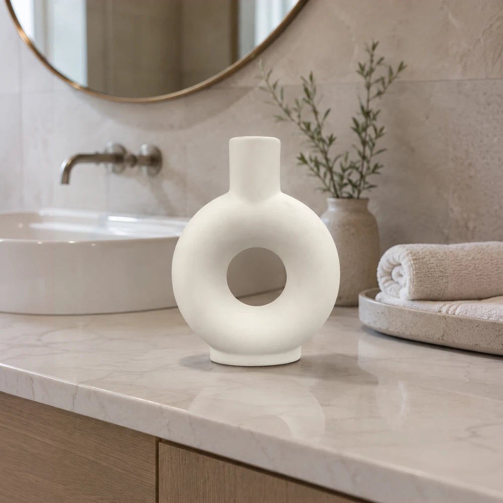 Minimalist white ceramic donut vase with a circular hollow center and a slim neck, placed on a marble bathroom counter.