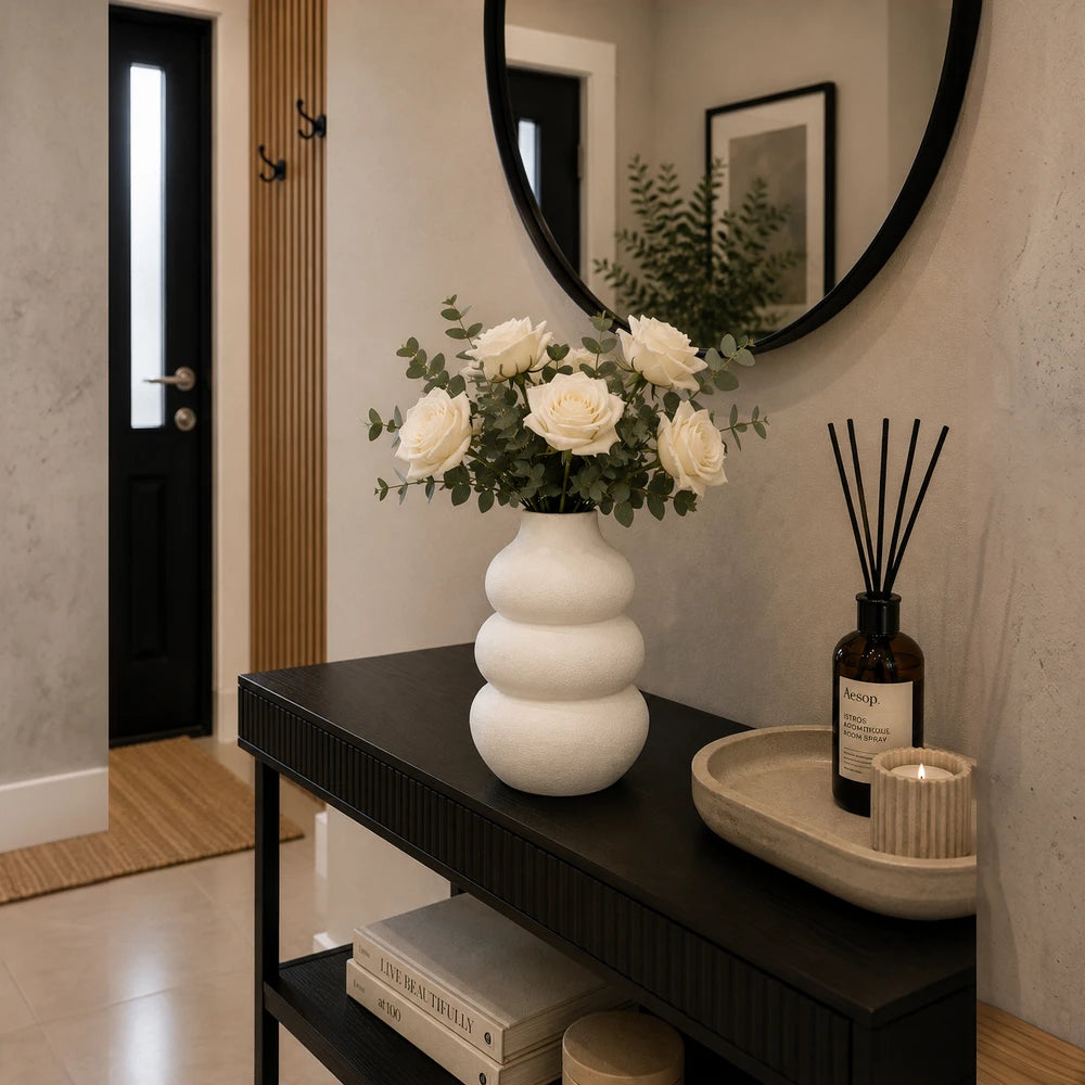 Modern white ceramic stacked pebble vase with white roses and eucalyptus on a dark console table.