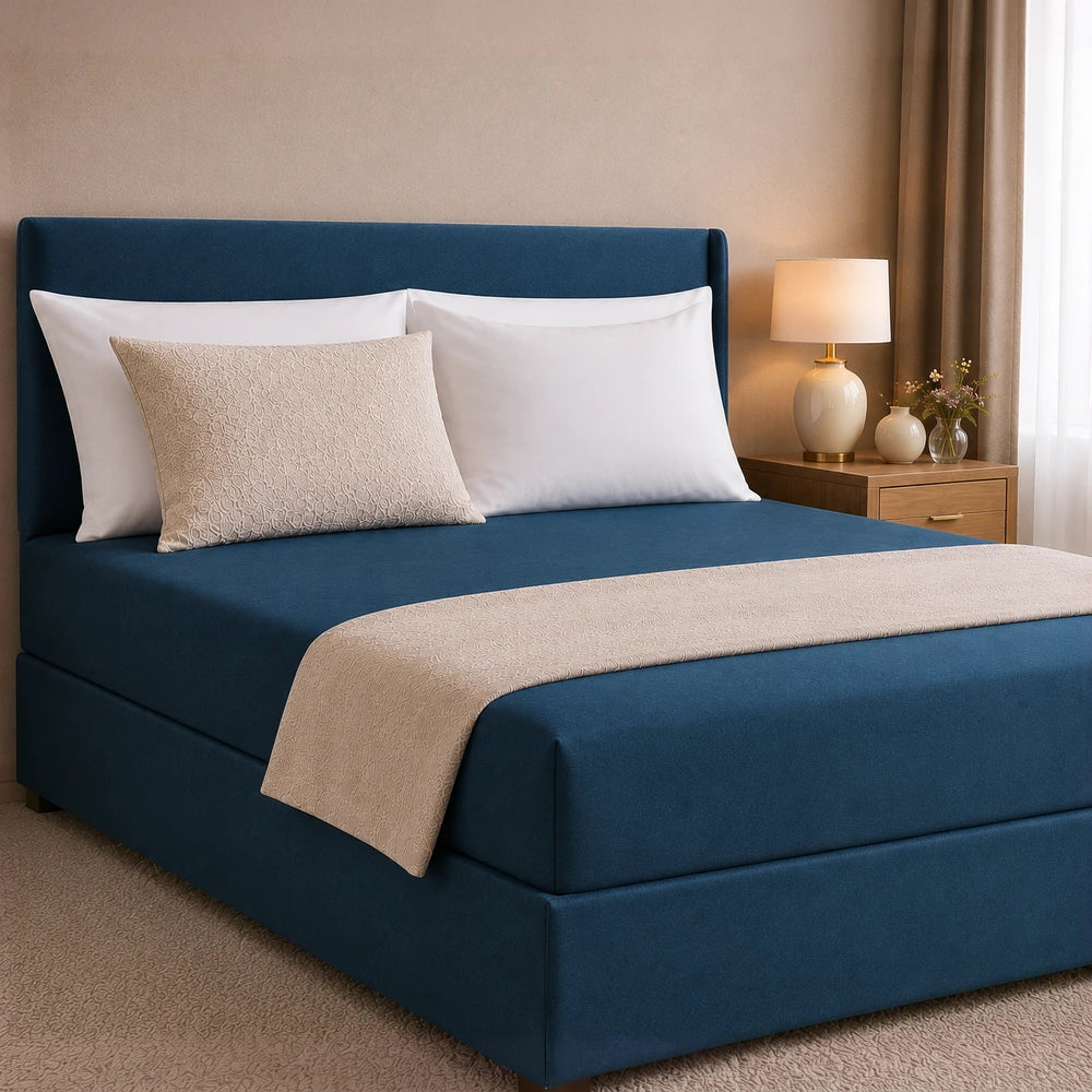 All-in-one Fitted Sheet and Base Cover in navy blue with a beige quilted throw, shown on a modern upholstered bed.