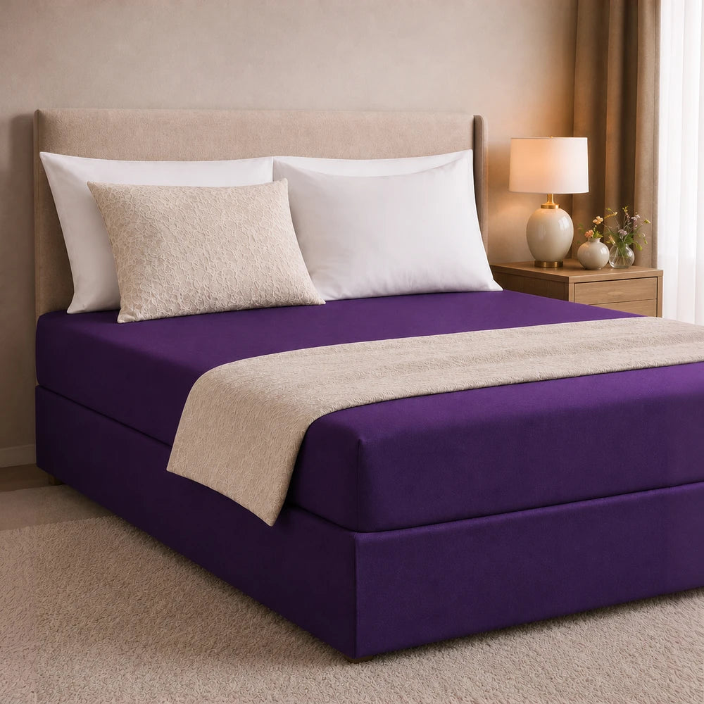 All-in-one fitted sheet and base cover in deep purple with a beige patterned top panel, shown on a neatly made bed.