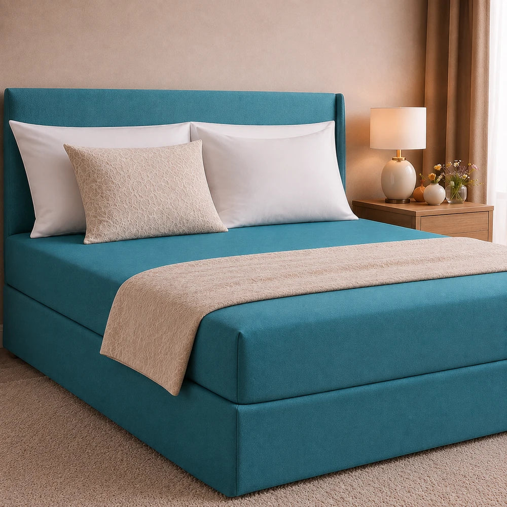 All-in-one fitted sheet and base cover in teal on a modern upholstered bed with white pillows and a beige decorative throw.